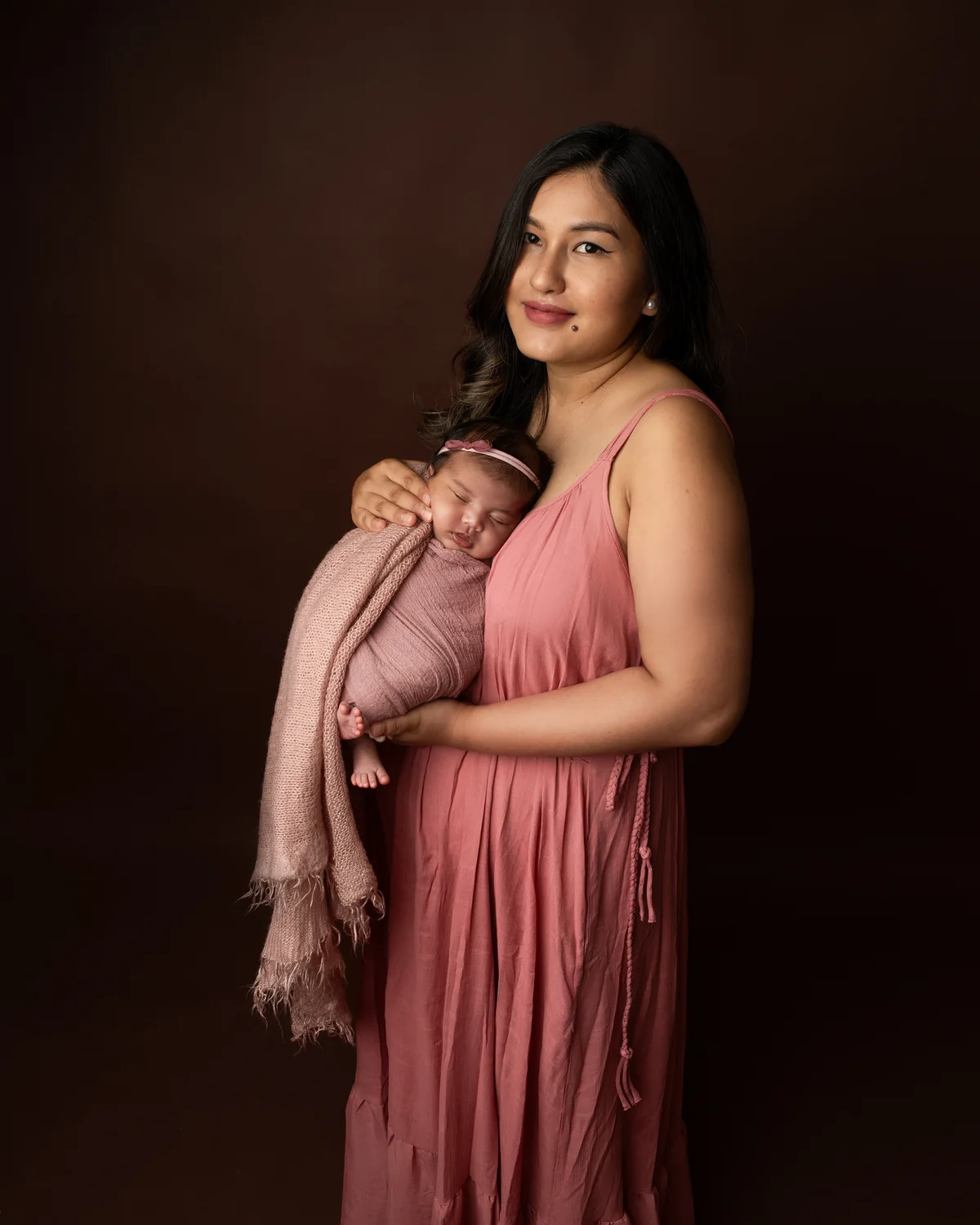 Woman holding a sleeping newborn in a warm rose-colored studio portrait