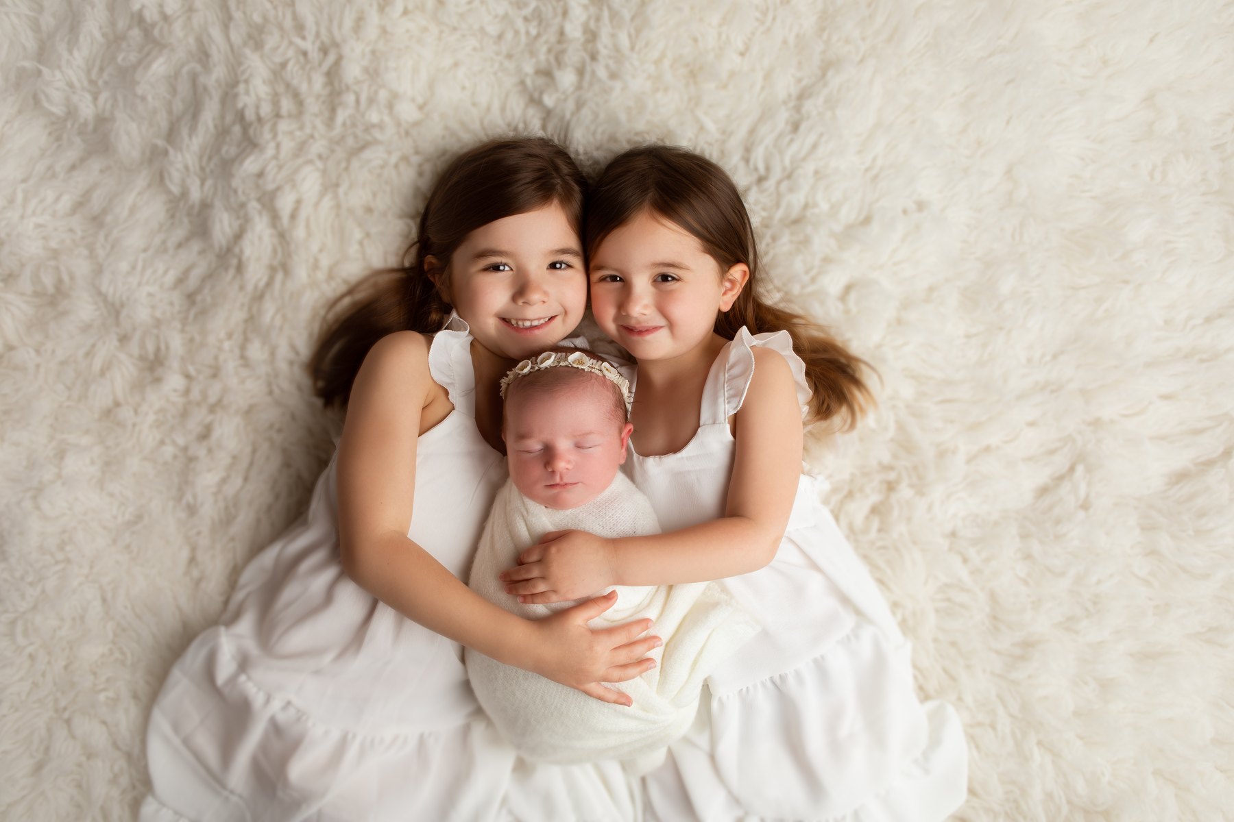 Two young sisters cuddling their sleeping newborn sibling on a soft cream blanket