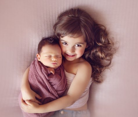 Little girl hugging swaddled newborn on pink background