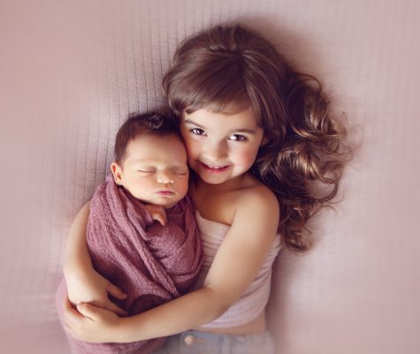 Little girl hugging swaddled newborn on pink background