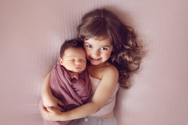 Little girl hugging swaddled newborn on pink background