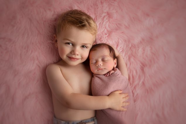 Toddler hugging swaddled newborn on pink blanket